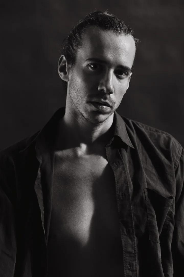 Zeyan Castracane. Head tilted in an open black shirt. Black-and-white portrait.