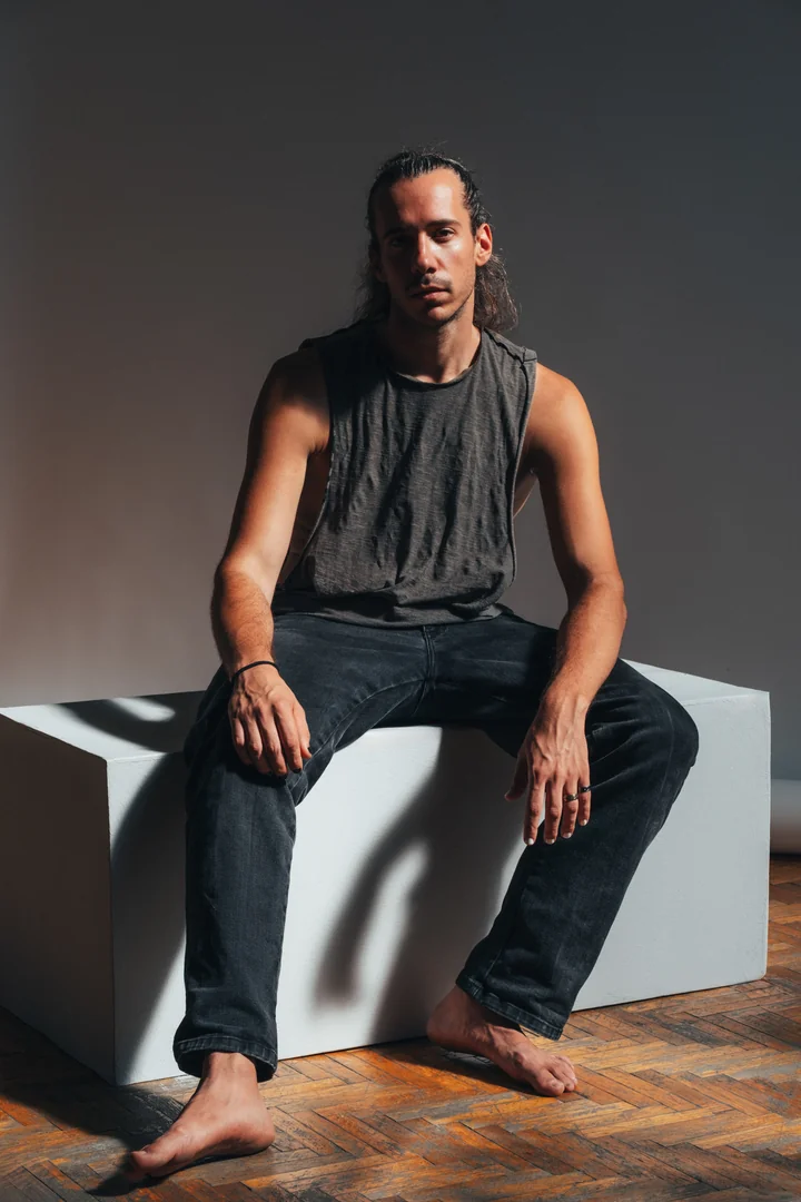 Zeyan Castracane. Seated barefoot on a white studio block. Full-length colour portrait.