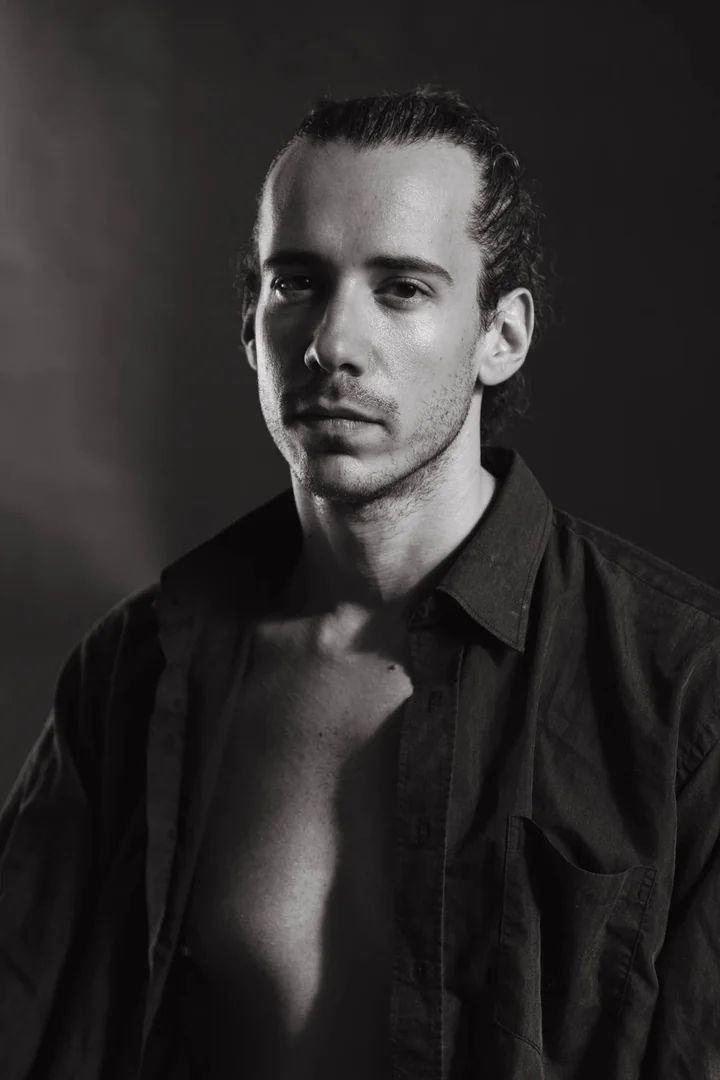 Zeyan Castracane. Facing the camera in an open black shirt. Black-and-white portrait.